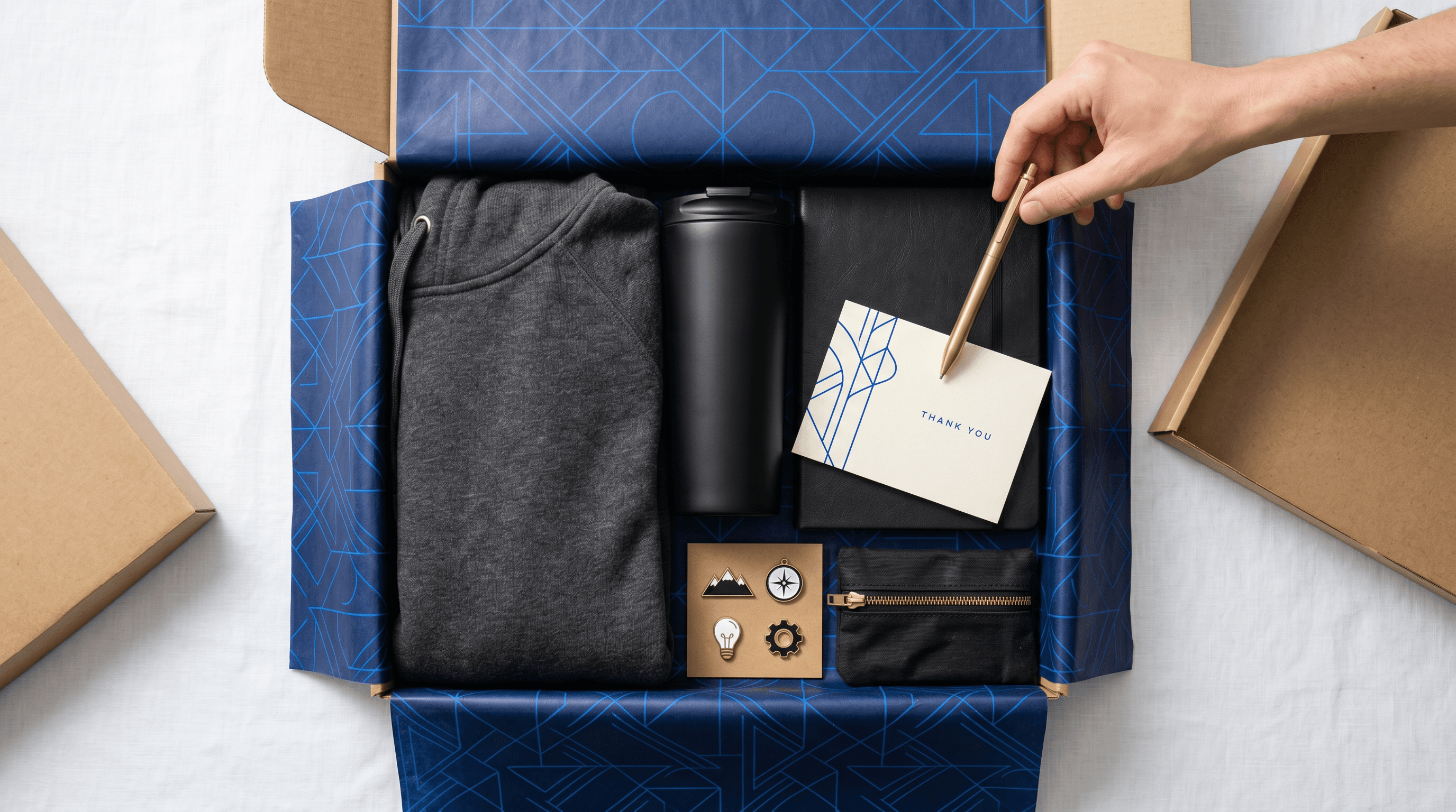 Overhead flat-lay of a custom branded swag box being assembled with hoodie, tumbler, journal, pen, pin set, and thank-you card on navy patterned tissue paper.